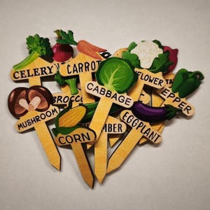 Op de afbeelding: Houten tuinmarkeringen met groente-illustraties en labels. De markeringen omvatten selderij, wortel, kool, paddenstoel, maïs, komkommer, aubergine, paprika en bloemkool. De labels zijn in een handschriftstijl.