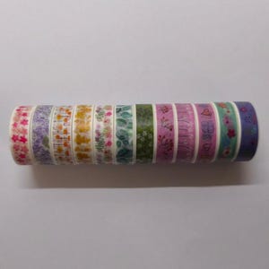 Puede incluir: Un rollo de cinta adhesiva washi decorativa con varios diseños florales. La cinta incluye patrones con flores y hojas rosas, moradas, amarillas, verdes y azules. Ideal para scrapbooking, manualidades y añadir un toque decorativo a los proyectos.