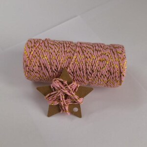 Può includere: Una bobina di spago rosa e oro con un'etichetta a forma di stella. Lo spago è di colore rosa chiaro con fili metallici dorati intrecciati. L'etichetta a stella è di cartone marrone, con spago avvolto.