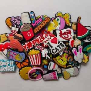 Puede incluir: Una colección de coloridos dijes para zapatos con varios diseños, incluyendo alimentos como palomitas de maíz y pizza, junto con frases como "Keep it Weird" e "I Love Cats". Los dijes son de varias formas y tamaños.