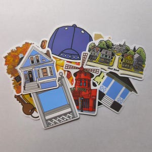 Op de afbeelding: Een verzameling kleurrijke stickers met diverse illustraties. Ontwerpen omvatten een blauwe baseballpet, een rode windmolen, huizen en een blauw huis met een sleutel. De stickers staan op een witte achtergrond.