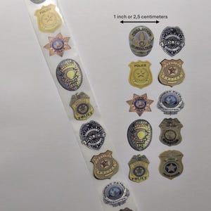 Pode incluir: Uma tira de autocolantes com vários desenhos de distintivos da polícia. Os distintivos são em ouro, prata e azul, com textos como "POLICE OFFICER" e "HIGHWAY PATROL". A imagem inclui um indicador de medida de 2,5 centímetros.