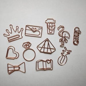 Könnte beinhalten: Eine Sammlung von roségoldenen Metall-Büroklammern in verschiedenen Formen, darunter eine Krone, ein Umschlag mit Herz, ein Kaffeebecher, ein Flamingo, das Wort love, ein Herz, ein Ring, ein geometrischer Diamant, eine Ananas, eine Schleife und ein Buch.