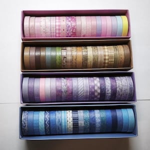 Op de afbeelding: Vier rechthoekige dozen met rijen decoratieve plakbandrollen. De bovenste doos bevat roze en pastelkleurige rollen, de tweede bevat aardetinten en bruine rollen, de derde bevat paarse tinten en de onderste doos bevat blauwe rollen.