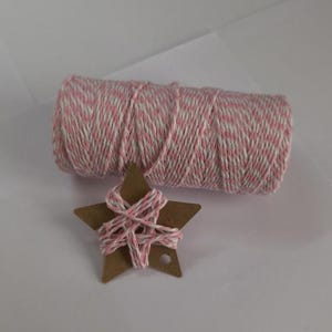 Puede incluir: Un carrete de hilo rosa y blanco se encuentra junto a una estrella de cartón marrón envuelta con el mismo hilo. El hilo está bien enrollado y la estrella tiene un pequeño agujero. Los artículos están sobre una superficie blanca.