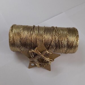 Pode incluir: Um carretel de fio metálico dourado com um enfeite de cartão em forma de estrela. O fio é enrolado firmemente e a estrela é enrolada com o mesmo fio dourado. Os itens estão em um fundo branco.