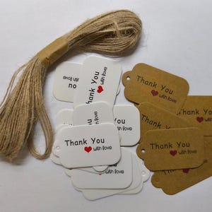 Può includere: Un insieme di etichette regalo bianche e marroni con la scritta "Thank You with love" e un cuore rosso. È incluso anche un rotolo di spago naturale, perfetto per attaccare le etichette.