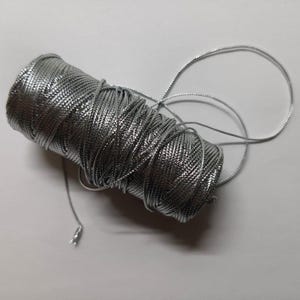 Pode incluir: Um carretel de cordão metálico prateado, bem enrolado, com um aspeto brilhante e texturizado. Um pedaço do cordão estende-se do carretel, com uma pequena etiqueta prateada presa. Adequado para artesanato e embrulho de presentes.