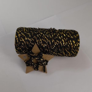 May include: A roll of black and gold twine rests on a brown cardboard star. The twine is tightly wound, with gold metallic threads woven through the black strands. The star is also wrapped with the same twine.