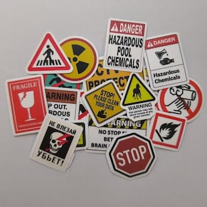 Op de afbeelding: Een verzameling diverse waarschuwingsstickers met duidelijke tekst en symbolen. De stickers tonen diverse ontwerpen, waaronder een voetgangersoversteekplaats, een stralingssymbool en het woord "GEVAAR" met verschillende gevarenwaarschuwingen.
