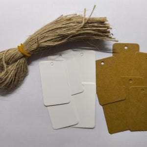 Può includere: Un fascio di spago con un elastico giallo, accanto a una collezione di etichette rettangolari. Alcune etichette sono bianche, altre marroni. Le etichette hanno un piccolo foro nella parte superiore per l'infilatura.