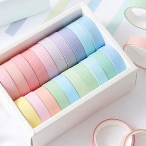 Op de afbeelding: Een witte doos gevuld met rollen pastelkleurige washi tape. De kleuren zijn roze, geel, blauw, groen en paars. Losse rollen roze en groene tape zijn ook zichtbaar, wat een kleurrijke en georganiseerde presentatie creëert.