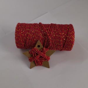 Könnte beinhalten: Eine Spule aus rotem Garn mit goldfarbenem Metallic-Faden und ein kleiner Holzstern, der mit demselben Garn umwickelt ist. Der Stern hat oben ein Loch. Die Artikel befinden sich auf einer weißen Oberfläche.
