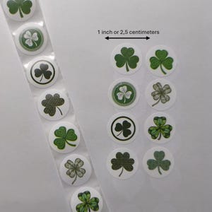 Op de afbeelding: Een strook ronde stickers met verschillende groene klaverontwerpen. De tekst "1 inch of 2,5 centimeter" staat boven de stickers. De stickers zijn wit met groene klavers, sommige met een zwarte rand. De klavers hebben verschillende patronen.