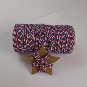Pode incluir: Um rolo de barbante vermelho, branco e azul está ao lado de uma estrela de papelão marrom com barbante enrolado. O barbante tem um esquema de cores patriótico, perfeito para artesanato ou embrulho de presentes. A estrela tem um pequeno furo para pendurar.
