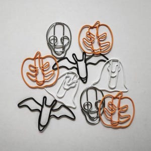 May include: A collection of Halloween-themed metal paper clips in various shapes and colours. The set includes orange pumpkin-shaped clips, white ghost-shaped clips, black bat-shaped clips, and silver skull-shaped clips, all arranged together.