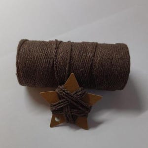 Könnte beinhalten: Eine Rolle braunes Garn und ein kleines, braunes sternförmiges Etikett, das mit dem gleichen Garn umwickelt ist. Das Garn und das Etikett sind vor einem weißen Hintergrund platziert. Der Stern hat ein kleines Loch unten.