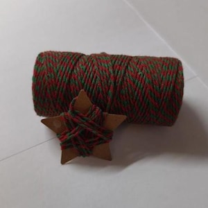 Pode incluir: Um rolo de barbante vermelho e verde e uma estrela de madeira enrolada no mesmo barbante. O barbante é uma combinação festiva de vermelho e verde, perfeito para artesanato e decorações de Natal. A estrela é de cor de madeira natural.