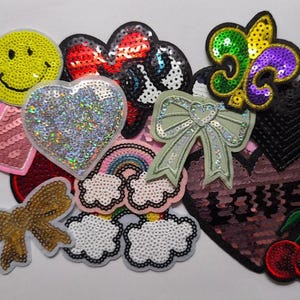 May include: A collection of colorful sequin patches in various shapes, including hearts, bows, a smiley face, a fleur-de-lis, clouds, a rainbow, and cherries. The patches are in shades of pink, red, green, gold, and silver. Some patches have the word "LOVE".