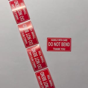 May include: Several red rectangular stickers with white text, reading "HANDLE WITH CARE", "DO NOT BEND", and "THANK YOU". The stickers are displayed on a white surface.