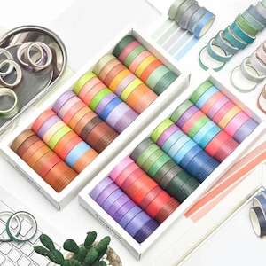 May include: Two white rectangular boxes filled with rows of colorful washi tape rolls in various shades including pink, blue, green, orange, and purple. Some rolls feature solid colors while others have a subtle grid pattern. Additional loose rolls are scattered nearby.