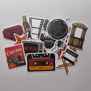 Può includere: Una collezione di adesivi a tema cinematografico. Include una bobina di film, una sedia da regista, un televisore vintage con il logo Netflix, una cassetta, un biglietto del cinema e una bottiglia di cola.