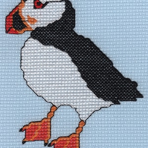 May include: A white and black cross-stitch of a puffin with orange feet and a red beak. The puffin is facing left and has a black eye.