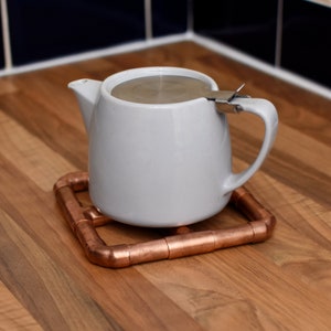 May include: A white teapot with a stainless steel lid and a copper pipe tea cozy.