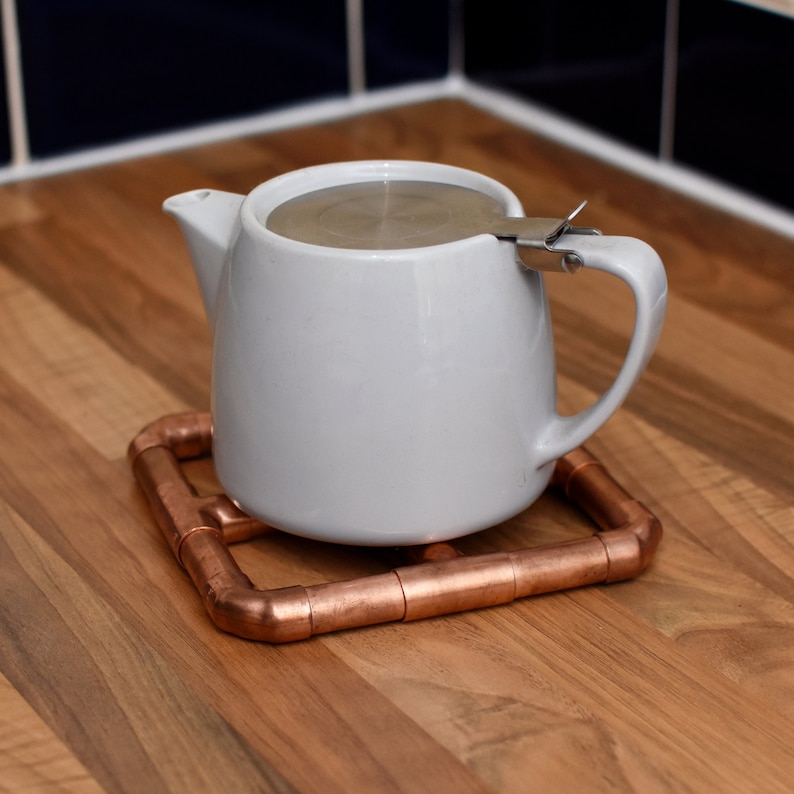 May include: A white teapot with a stainless steel lid and a copper pipe tea cozy.