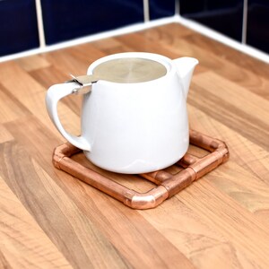 May include: A white teapot with a stainless steel lid sits on a copper pipe coaster. The coaster is on a wood surface.