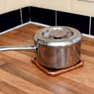 May include: A stainless steel pot with a lid sits on a copper pipe trivet on a wood countertop.