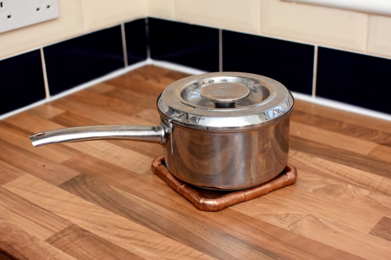 May include: A stainless steel pot with a lid sits on a copper pipe trivet on a wood countertop.