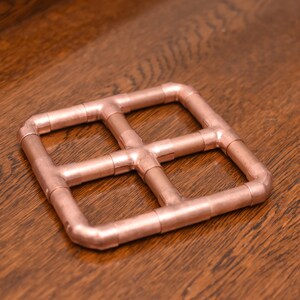 May include: A copper-colored metal coaster with a square shape and a grid pattern. The coaster is made of four pieces of pipe that are connected at the corners.