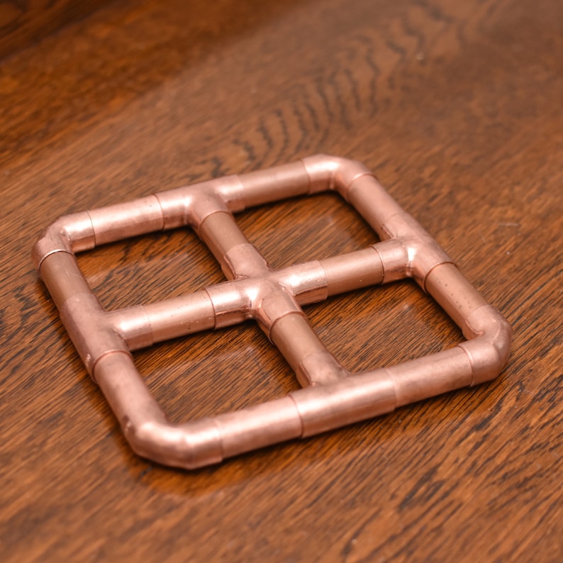 May include: A copper-colored metal coaster with a square shape and a grid pattern. The coaster is made of four pieces of pipe that are connected at the corners.
