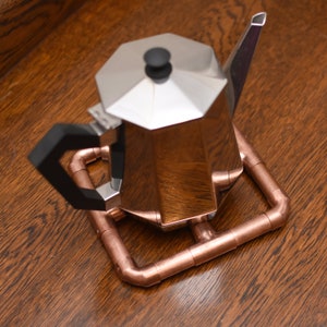 May include: A silver coffee pot with a black handle sits on a copper pipe trivet. The trivet is made of copper pipes that have been cut and joined together to form a square shape.