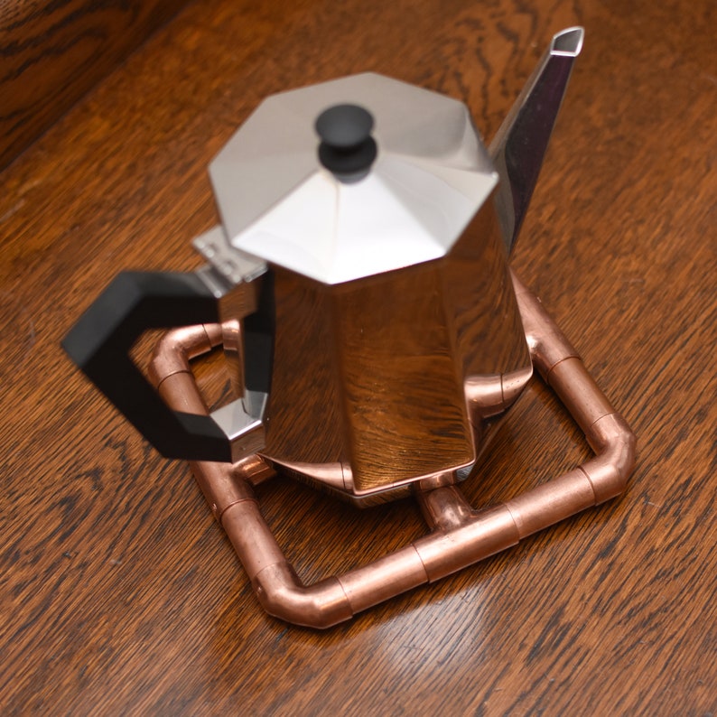 May include: A silver coffee pot with a black handle sits on a copper pipe trivet. The trivet is made of copper pipes that have been cut and joined together to form a square shape.