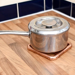 May include: A silver pot with a lid sits on a copper pot holder. The pot holder is on a wooden surface.