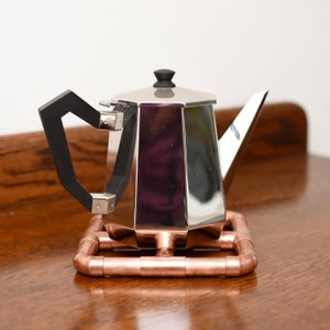 May include: A silver teapot with a black handle sits on a copper pipe trivet. The teapot has a geometric shape and a shiny finish.