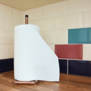 May include: A copper paper towel holder with a white paper towel roll. The holder is on a wooden worktop in front of a tiled wall with blue, green, and red tiles.
