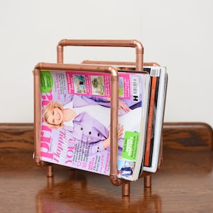 May include: A copper magazine rack with a modern design. The rack is holding several magazines, including one with the title "Happy You!" on the cover.
