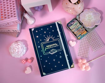 May include: A navy blue Reading Journal with a sun and star design, a silver elastic closure, and gold corner protectors. The journal is surrounded by decorative items on a pink surface, including a stack of pink journals.