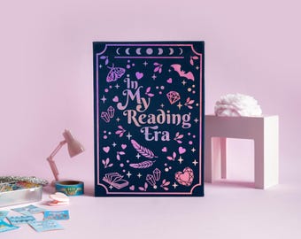 May include: A dark blue book with the text "In My Reading Era" in a decorative font, surrounded by illustrations of moons, bats, crystals, and hearts. The book is on a pink background.