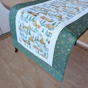 May include: A quilted table runner featuring a repeating pattern of vintage cars, cabins, and evergreen trees on a white background. The runner is bordered with a dark green fabric adorned with gold snowflakes. The runner is placed on a wooden table.