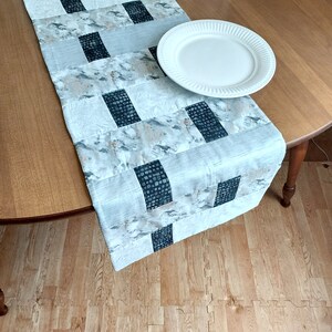 May include: A white table runner with a black and gray geometric pattern. The runner is on a wooden table with a white plate on top.