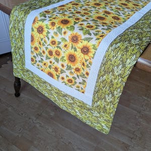 May include: A green and white table runner with a sunflower pattern. The runner has a white border with a green outer border.