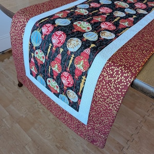 May include: A decorative table runner featuring a black background with a pattern of red, white, and gold lanterns. The runner has a white border and a red border with gold square accents. The runner is placed on a wooden table.