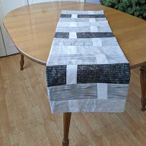 May include: A rectangular table runner with a patchwork design. The runner features alternating sections of gray, white, and black fabrics. The table runner is displayed on a wooden table.