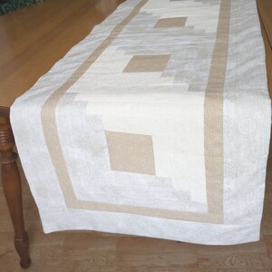 May include: A rectangular table runner with a quilted design. The runner features a central pattern of light beige and cream-colored squares and rectangles, bordered by a tan fabric with a subtle pattern. The overall aesthetic is neutral and rustic.