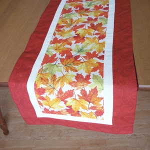 May include: A red table runner with a white border and a center panel featuring a pattern of autumn leaves in shades of red, orange, yellow, and green.
