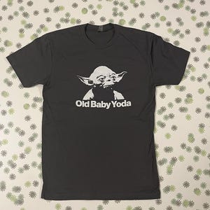 May include: A dark gray t-shirt with a white graphic of Yoda from Star Wars. The text "Old Baby Yoda" is printed below the image.